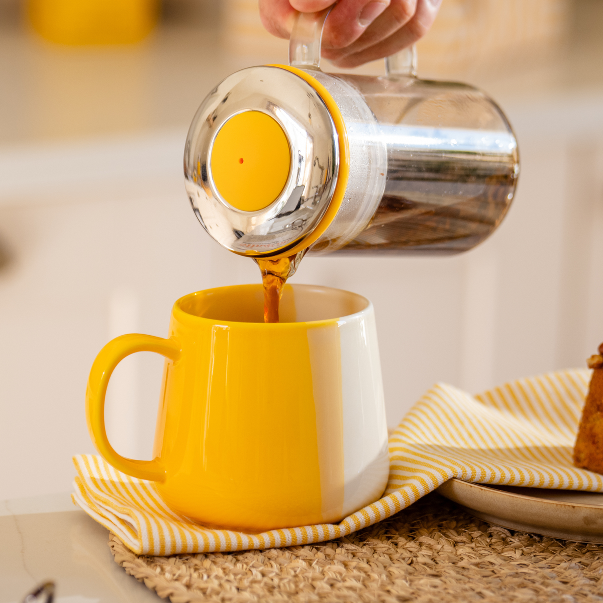 A hand pours hot english breakfast tea from a clear glass tea pot for one with a yellow lid into a yellow and white ceramic brew tea mug.