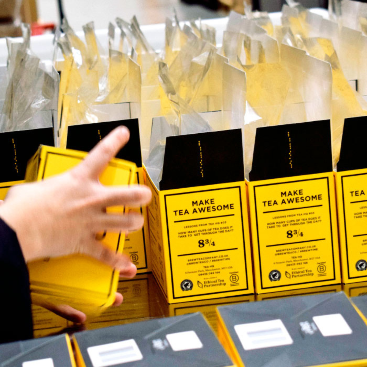 In warehouse hand holds a stack of bright yellow tea bags packing tea to bags, with multiple matching yellow and black tea bag boxes arranged on a table. The front box reads 'MAKE TEA AWESOME'.