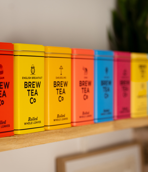 A row of colorful Brew Tea Co. tea tins in a variety of colours designed to hold loose leaf tea arranged on a light wooden shelf against a softly blurred indoor background.