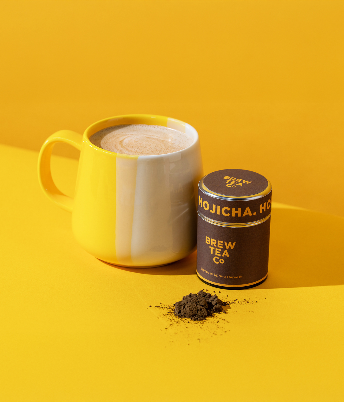 A yellow and white ceramic mug filled with a frothy Hojicha Latte sits beside a tin of Brew Tea Company Hojicha