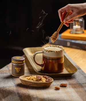 A hand holds a wooden honey dipper above a hot mug of a Hojicha Latte on a tray, with a small jar labeled Hojicha Brew Tea Co. and a small bowl of nuts.